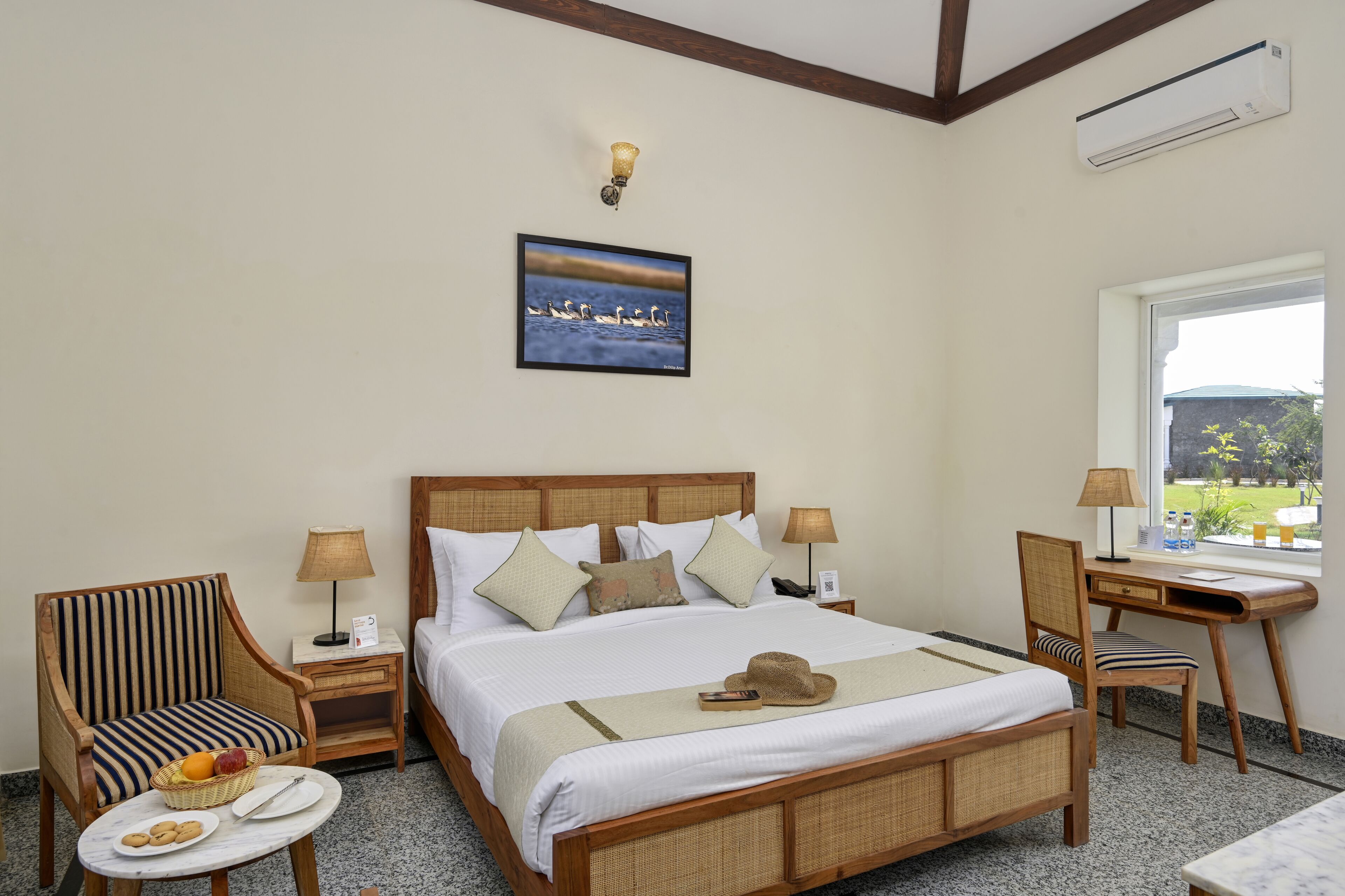 A hotel room with a bed, a desk, a chair, a small table with snacks, a lamp, and a picture of swans on the wall.