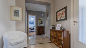 Un couloir avec un fauteuil blanc, des étagères en bois ornées d’objets décoratifs et des œuvres d’art encadrées accrochées aux murs.