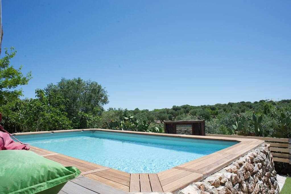 Una piscina rectangular amb un pont de fusta i un marc de pedra, envoltada de vegetació exuberant i un cel blau clar.
