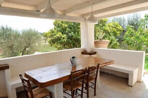 Mesa de madeira com cadeiras e um banco sob um alpendre coberto, com vista para um jardim com árvores e plantas.