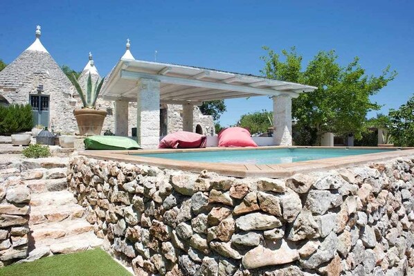 Uma piscina de pedra com pérgola, paredes de pedra e um edifício de pedra com telhados cônicos.