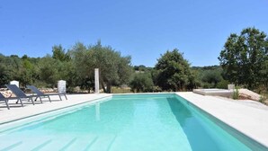 Una piscina rectangular con agua azul cristalina, rodeada por una terraza blanca. Hay sillas de descanso a lo largo de la piscina.