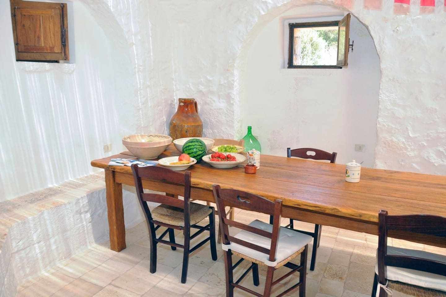 A rustic dining area with a wooden table and chairs, surrounded by white walls and a small window.