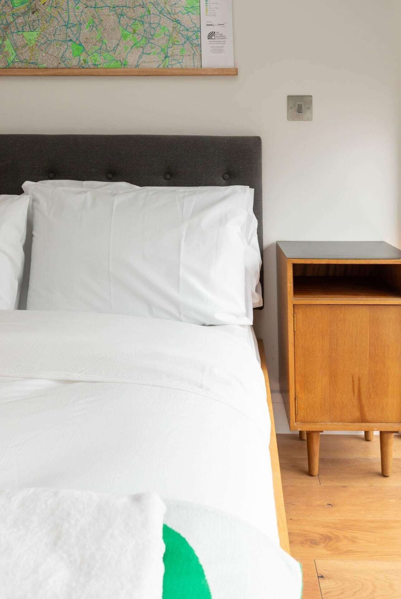 A neatly made bed with white bedding and a green accent, a grey upholstered headboard, and a wooden nightstand with a drawer.