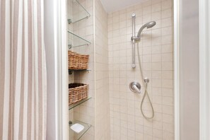 Une douche équipée d'un pommeau de douche à main et d'un robinet mural, d'étagères en verre avec des paniers en osier, et d'une paroi de douche en verre.