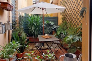 Une petite terrasse avec une table et des chaises, entourée de plantes en pot et d’une clôture en métal.