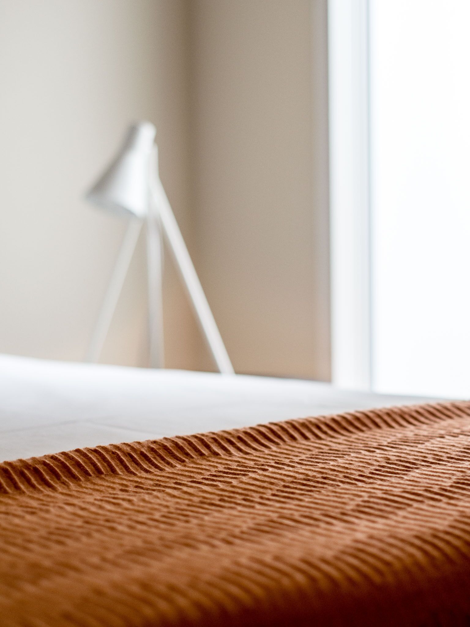 A close-up of a textured brown fabric, possibly a bedspread, with a blurred background featuring a white lamp and a window.