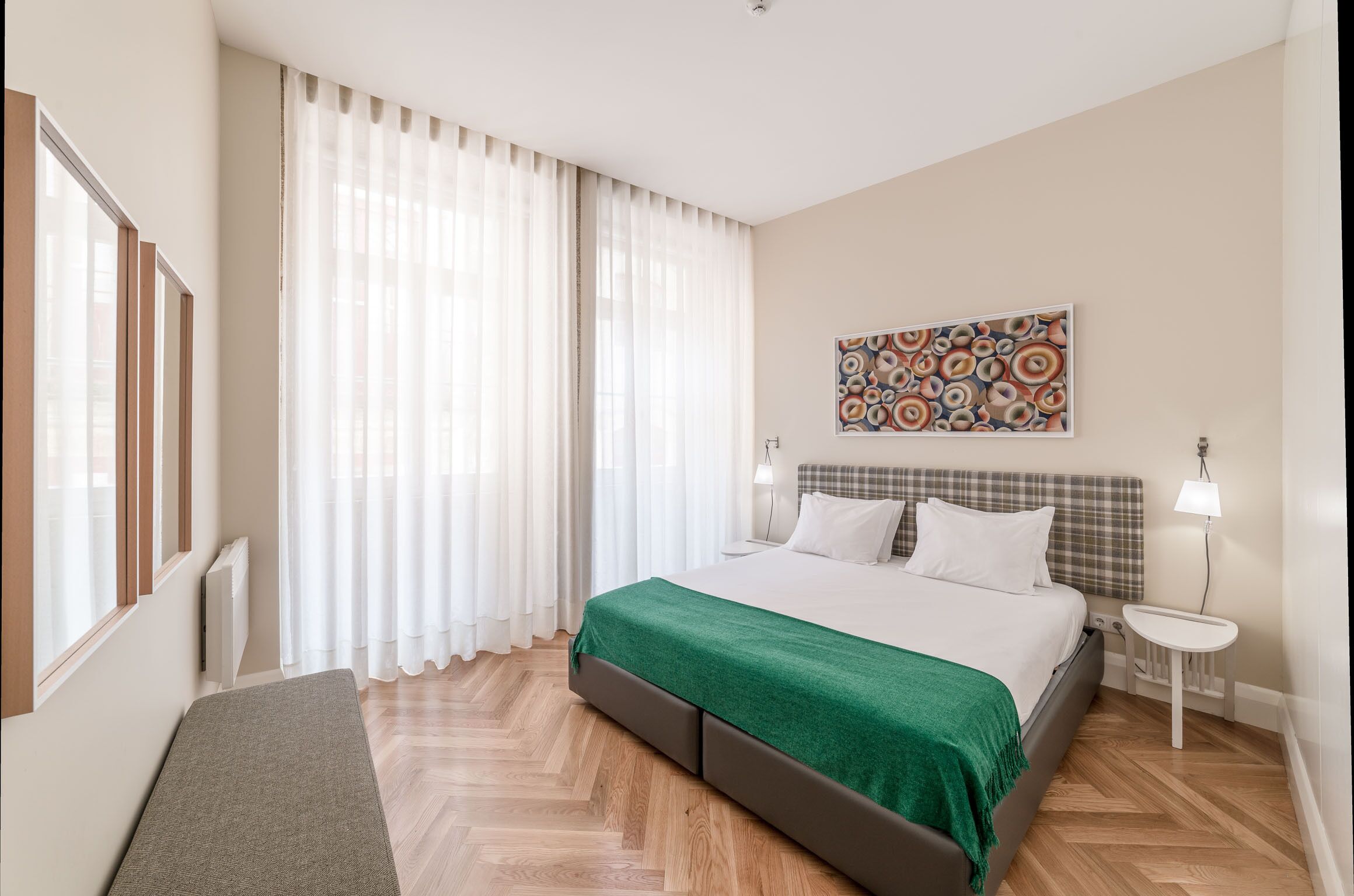 A modern hotel room with a large bed, a bench, a small table, and a window with sheer curtains.