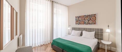 A modern hotel room with a large bed, a bench, a small table, and a window with sheer curtains.