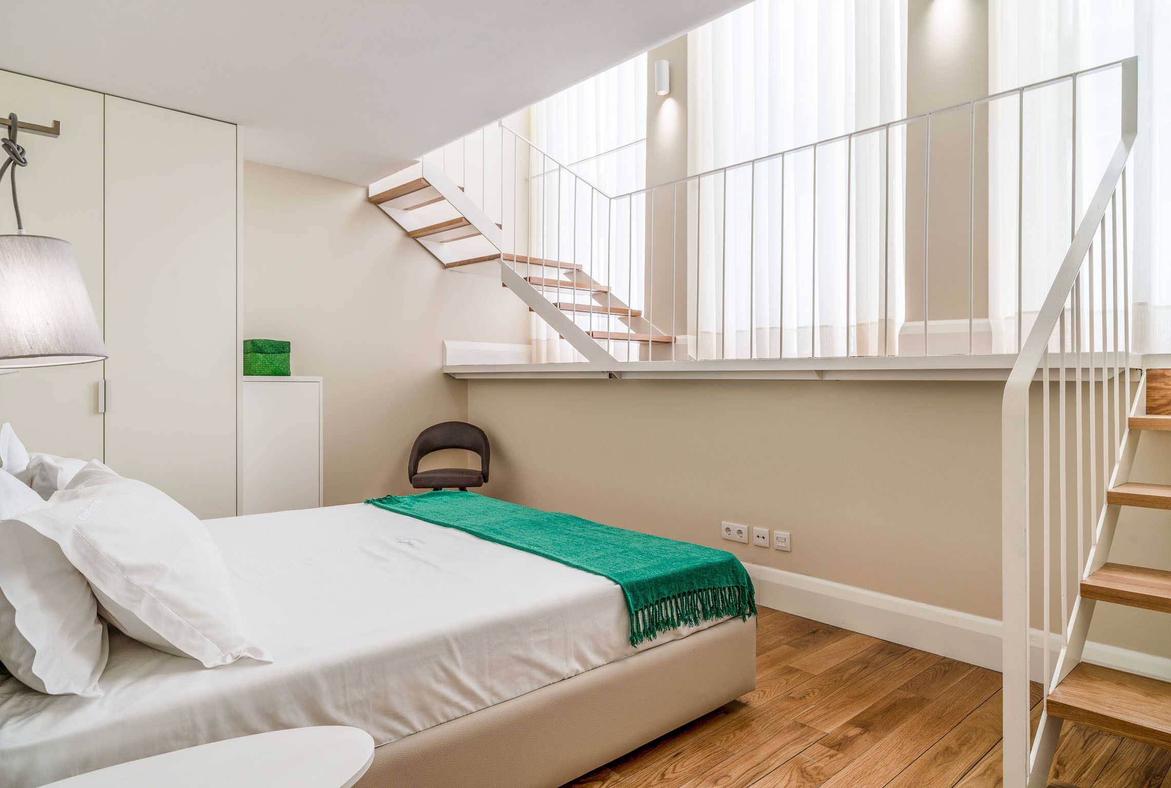 A bedroom with a bed, a green throw blanket, a chair, a desk, and a staircase.
