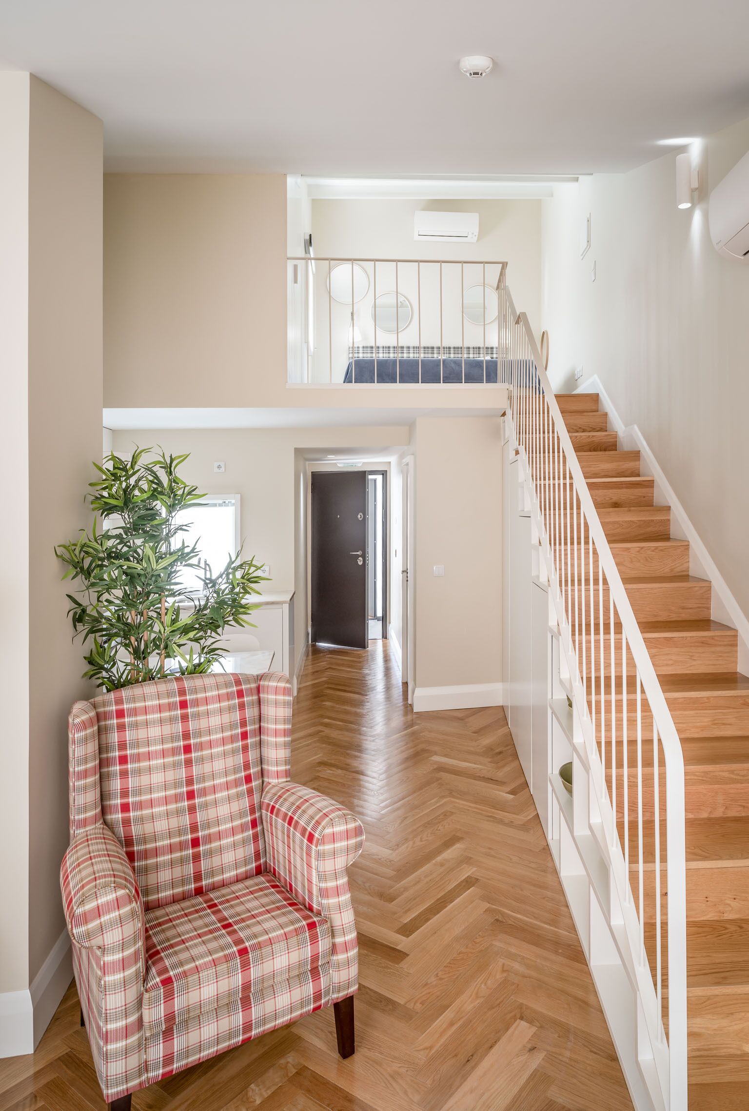 A spacious living area with a wooden staircase, a plaid armchair, and a large plant.