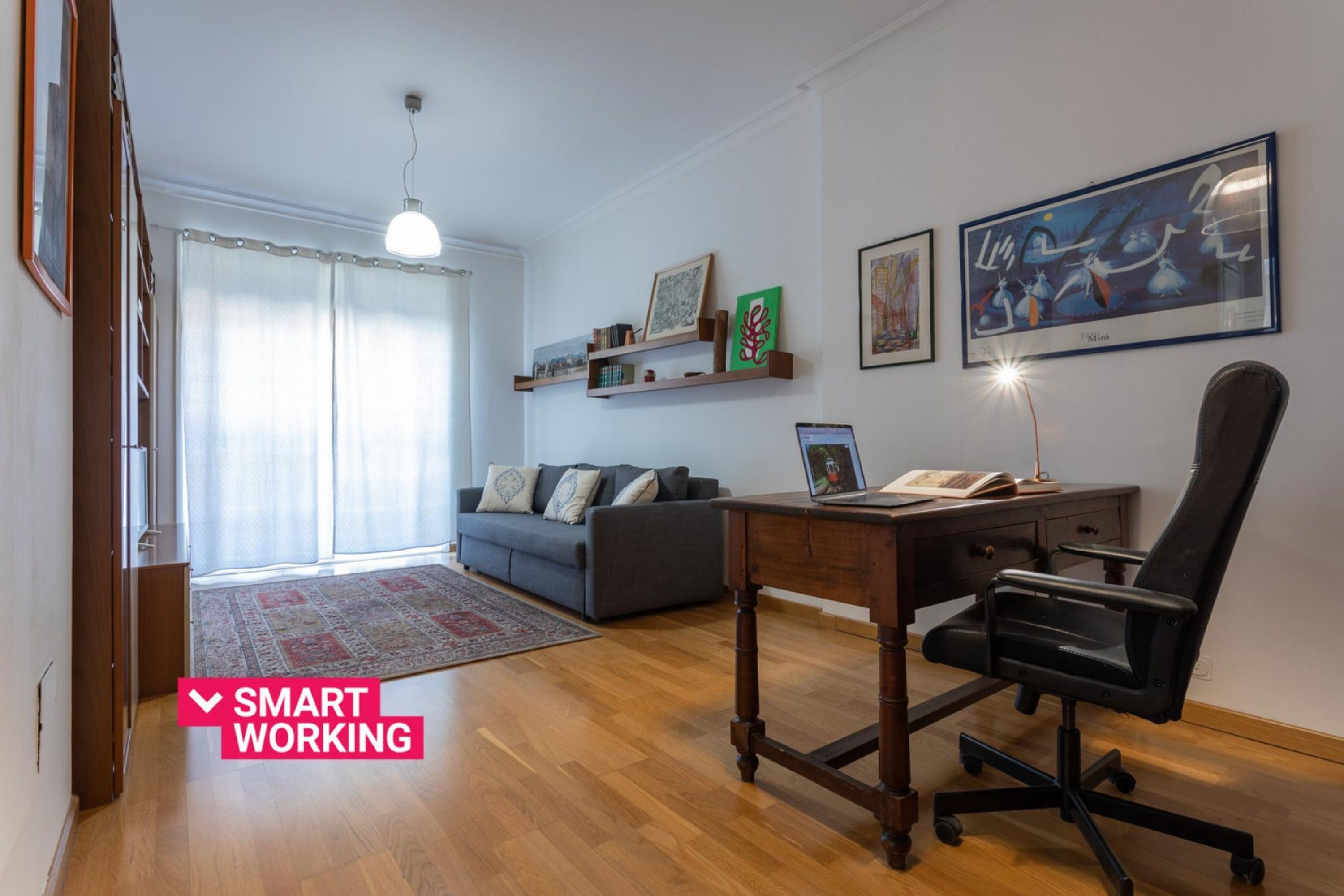 Ein modernes Wohnzimmer mit Holzboden, einem grauen Sofa, einem Schreibtisch mit Stuhl und Kunstwerken an den Wänden.