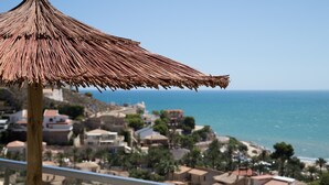 A balcony with a thatched umbrella, overlooking a coastal town with buildings and the sea.
