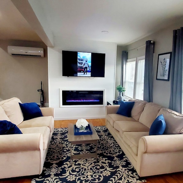 Una sala de estar con dos sofás beige, una mesa de centro con un jarrón, un televisor de pantalla plana sobre la chimenea y una alfombra estampada.
