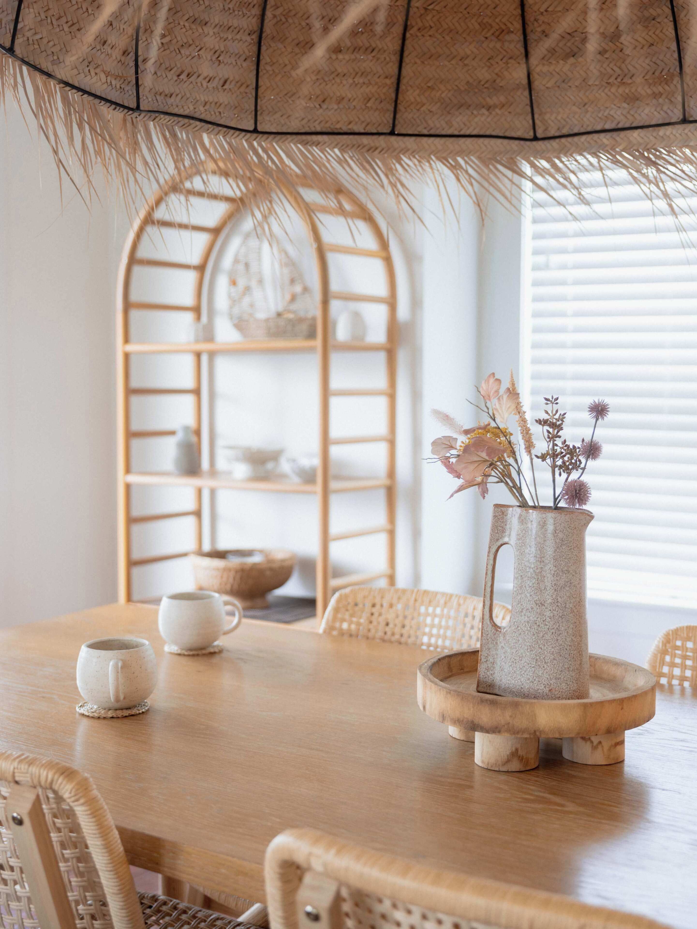 A wooden dining table with a woven basket, a ceramic pitcher with flowers, and a woven chair.