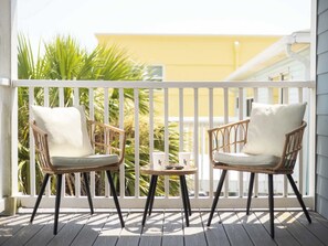 A balcony with two wicker chairs and a small table, overlooking a yellow building and green foliage.
