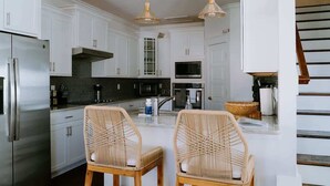 Una cocina moderna con armarios blancos, electrodomésticos de acero inoxidable, sillas de mimbre e iluminación colgante.