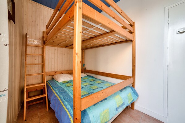 Una cama litera de madera con escalera, colcha azul y verde, y una puerta blanca al fondo.
