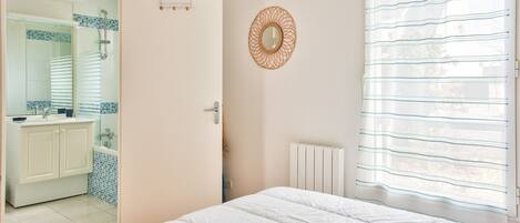 A bedroom with a large bed, a bathroom with a shower, and a view through a window with blinds.