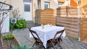 Een terras buiten met een tafel gedekt voor twee personen, omringd door houten stoelen, een houten poort en een gebouw met een bord.