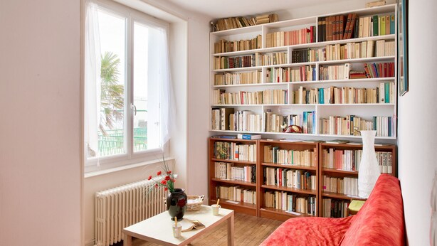 Een kamer met een grote boekenkast, een radiator, een tafel met een vaas en een rode bank.