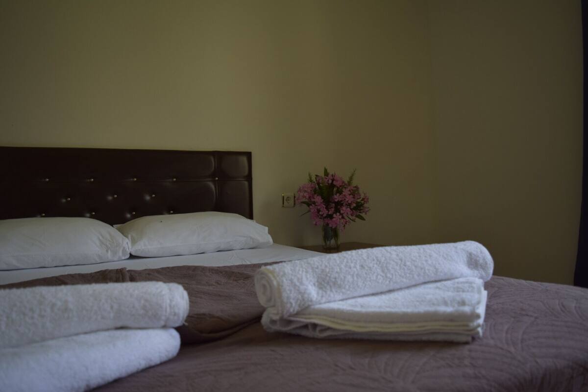 A hotel room with a large bed, two pillows, a vase with flowers, and folded towels on the bed.