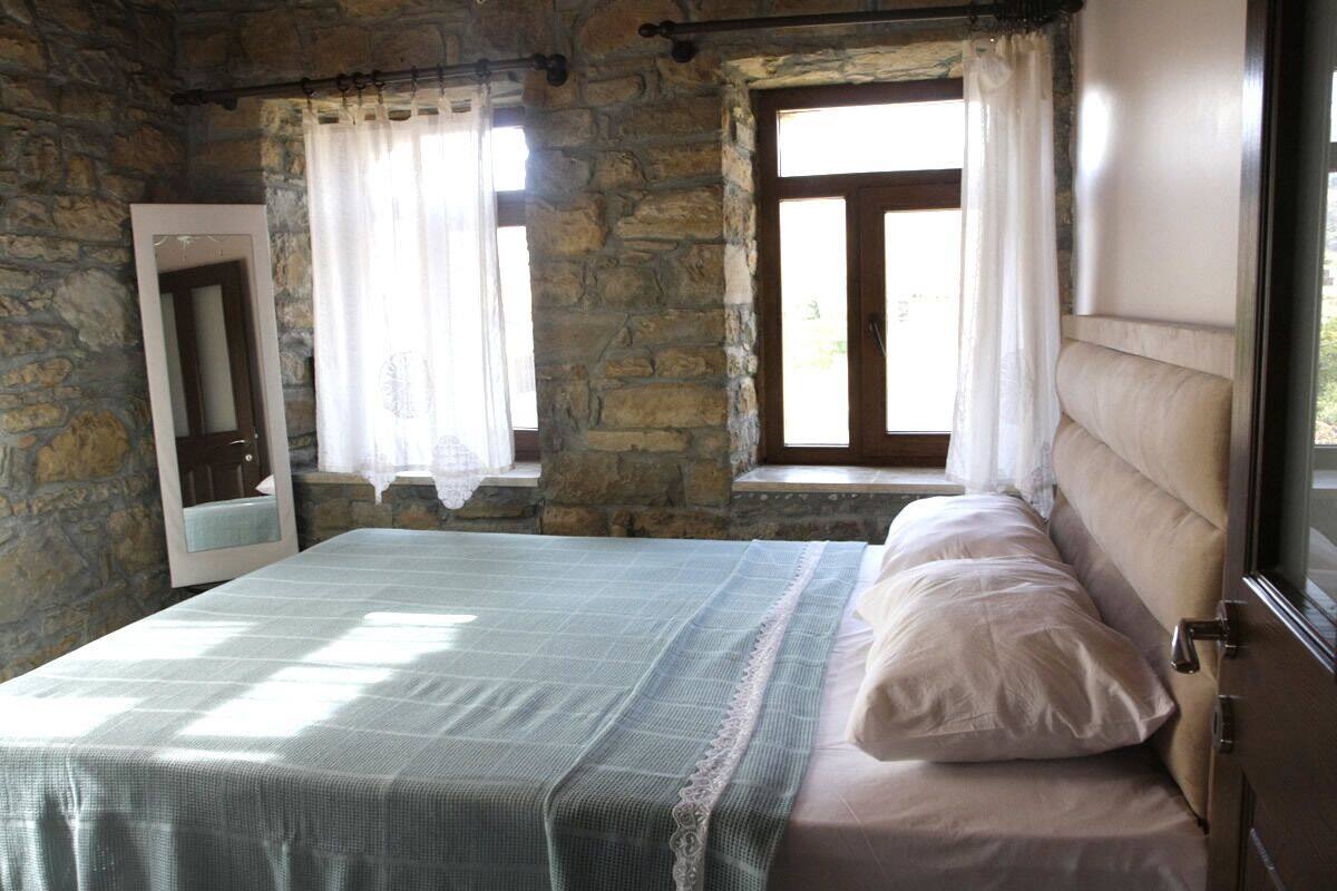 A bedroom with a stone wall, a bed with a white headboard, and two windows with curtains.