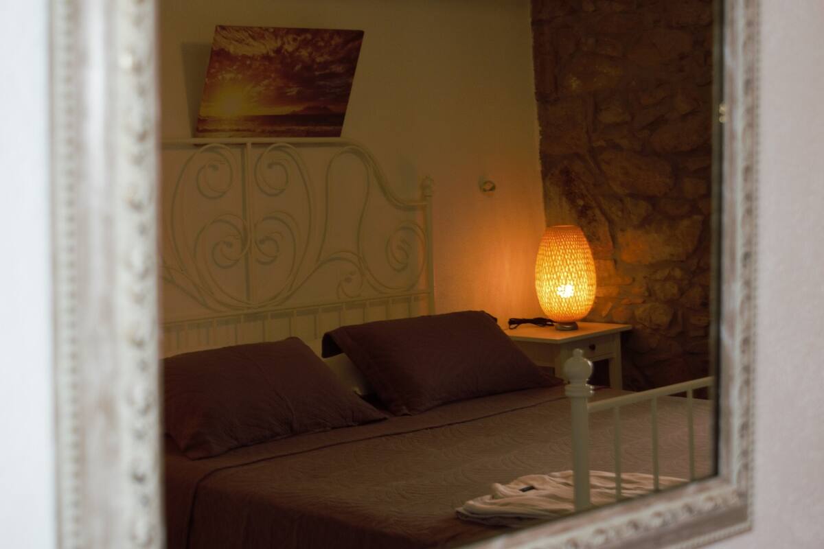 A bedroom with a bed, pillows, a nightstand with a lamp, a painting on the wall, and a stone wall.
