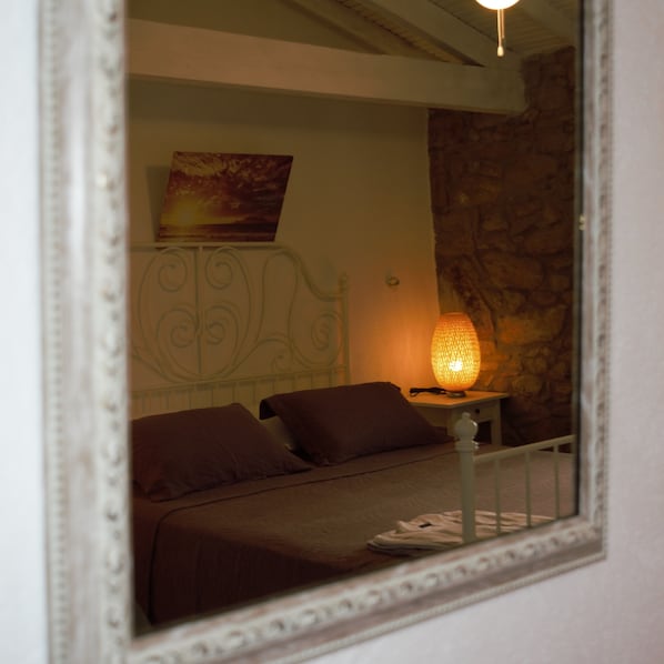A bedroom with a bed, pillows, a nightstand with a lamp, a painting on the wall, and a stone wall. - Kalyopi Butik Otel (Gökçeada)