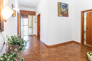 A hallway with hexagonal brown tiles, a wicker plant stand, and a painting on the wall.