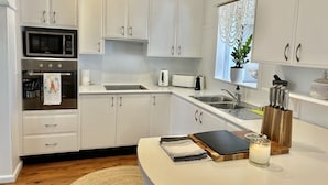 A modern kitchen with white cabinets, a microwave, an oven, a sink, and a window with a plant. - The Ultimate Family Friendly Getaway (Long Jetty)