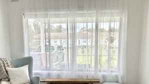 A living room with a light blue armchair, a bench, and a view of a residential area through sheer curtains. - The Ultimate Family Friendly Getaway (Long Jetty)