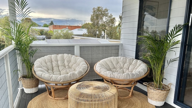 A balcony with two cushioned chairs and a woven ottoman, surrounded by potted plants. - The Ultimate Family Friendly Getaway (Long Jetty)