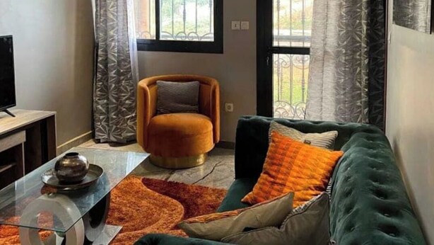Una sala de estar moderna con una mesa de centro de vidrio, un sofá verde y un sillón cómodo. Se aprecia un ventanal con celosías de hierro y vistas a la vegetación.