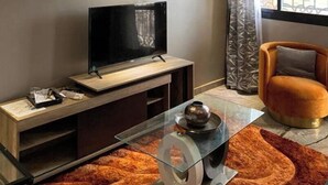 Una sala moderna con un televisor de pantalla plana sobre un soporte de madera, una mesa de centro de vidrio con un objeto decorativo, un tapete estampado y un sillón naranja cómodo.