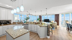 A modern kitchen with granite countertops, white cabinetry, and a large island. There is a dining area with a view of the ocean. - NEW RENTAL-Oceania 13-Luxury 4000 SQ FT Gulf Front Condo-Signature Properties (Gulf Shores)