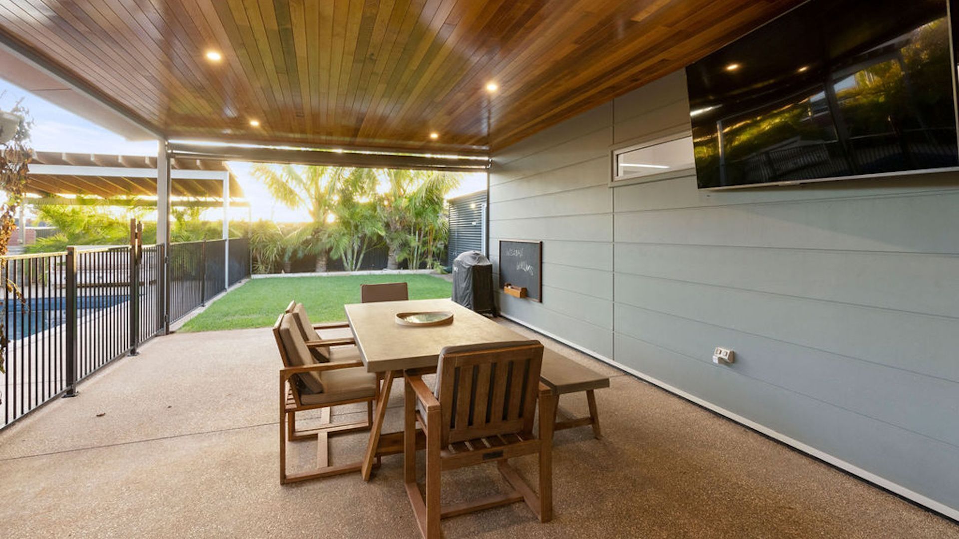 Un patio exterior con una mesa y sillas de madera, un televisor montado en la pared y vista al jardín trasero.