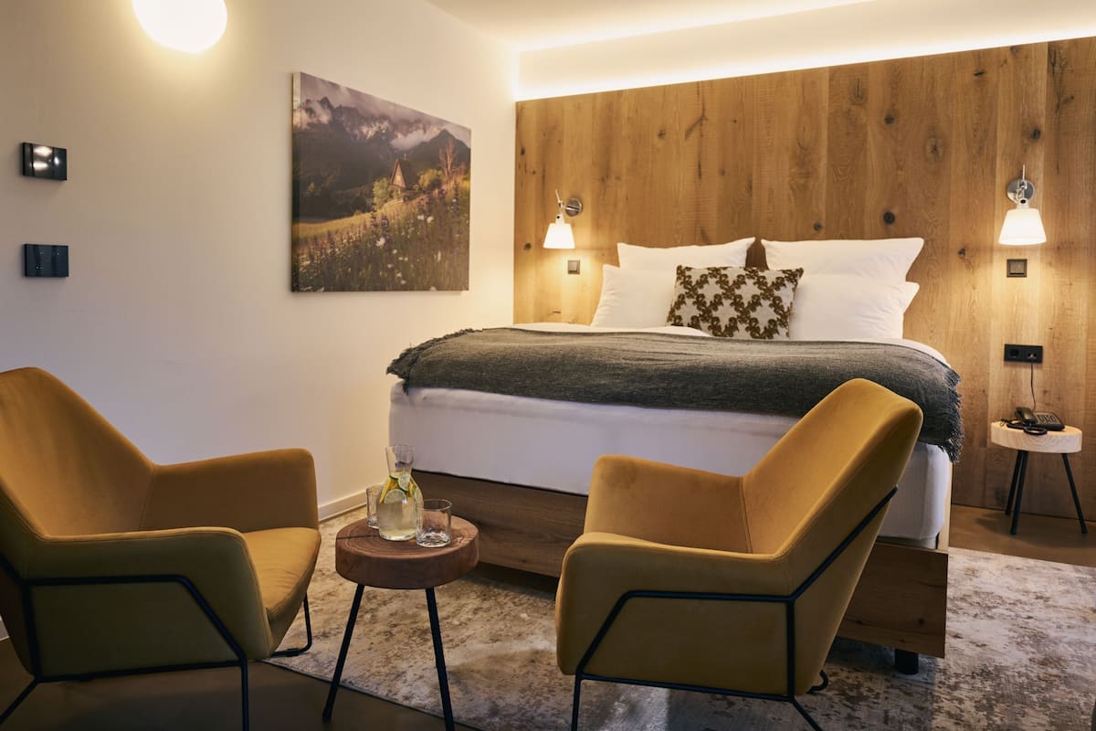 Een moderne slaapkamer met een houten hoofdbord, een groot bed met wit en donkergrijs beddengoed, twee gele fauteuils, een klein rond tafeltje met een fles en glas, en twee wandlampen.