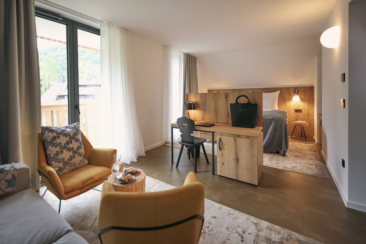Een moderne hotelkamer met een houten bureau, een comfortabele bank, een klein rond tafeltje met een glas, en uitzicht op het buitenlandschap door grote ramen.
