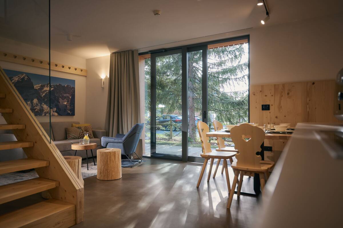 Een modern interieur met een houten trap, een eethoek met houten stoelen, een bank en een groot raam met uitzicht op bomen en een berg.