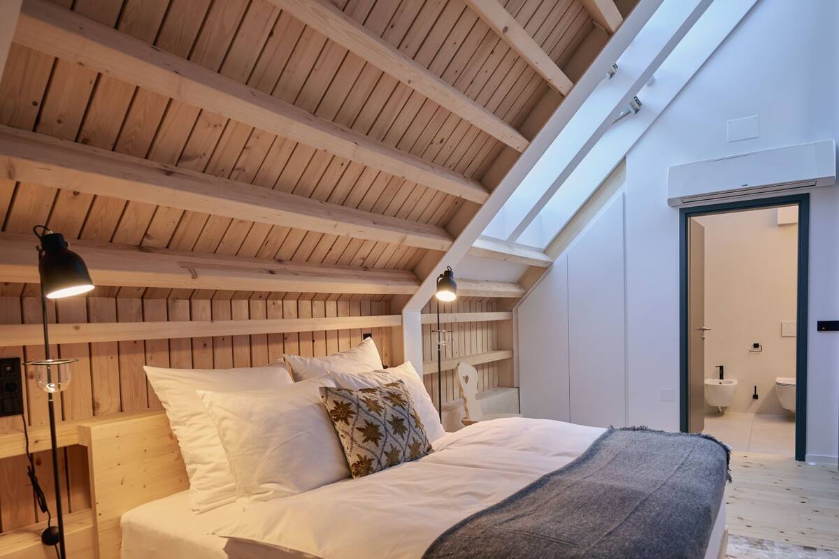 Een slaapkamer met schuine plafond, houten balken, een bed met witte beddengoed, een bedlampje en een klein houten krukje.
