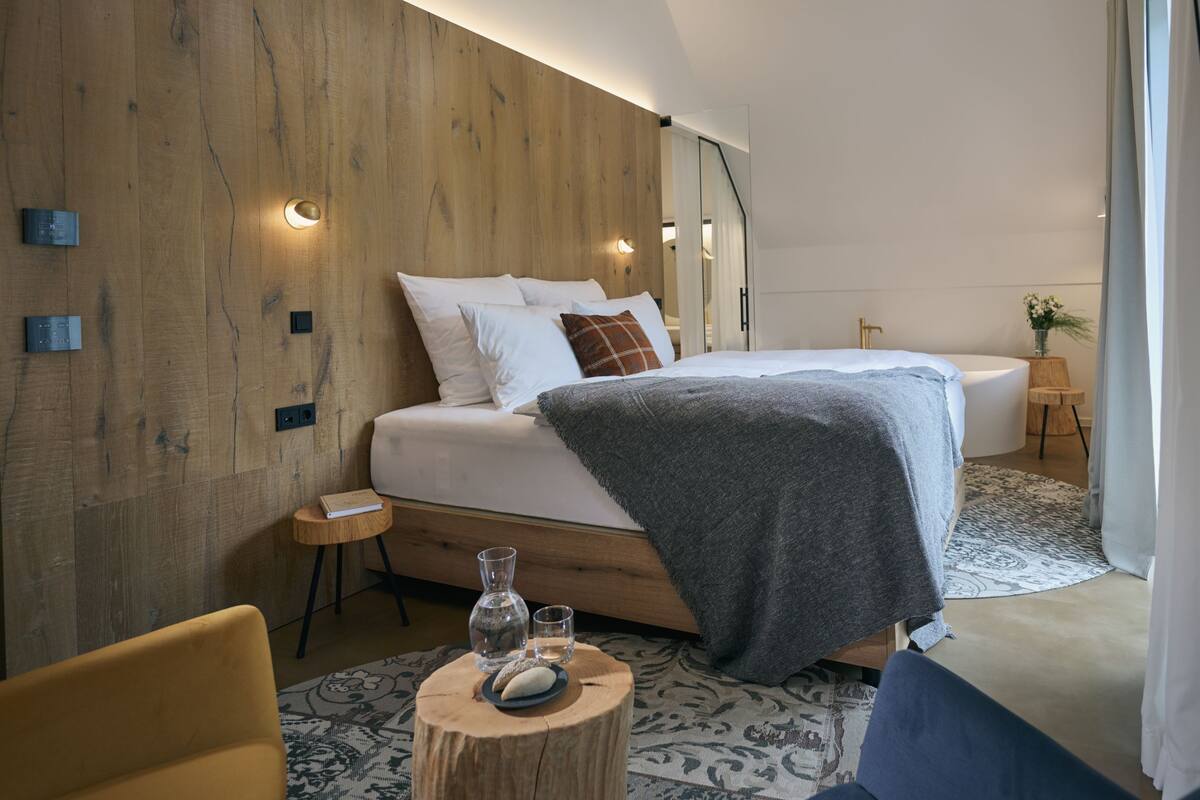 Een moderne slaapkamer met een houten hoofdbord, een groot bed met een grijze deken, nachtkastjes met lampen en een klein rond tafeltje met een vaas en glaasjes.
