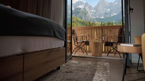 Ein Schlafzimmer mit einem Bett, einem Nachttisch aus Holz und Blick auf einen Balkon mit Gartenmöbeln und Bergblick.