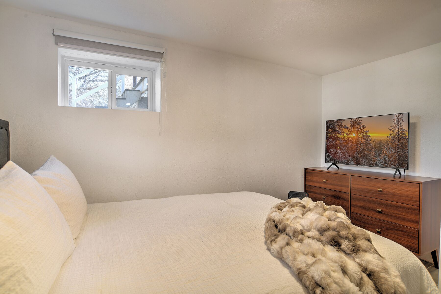 A bedroom with a bed, a wooden dresser, a television on the dresser, and a window with a view of trees.