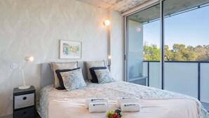 A modern bedroom with a large bed, two bedside tables, a nightstand, a framed picture on the wall, and a balcony with a view of trees. - Luxurious Soft Flat | Balcony and Pool by HostWise (Vila Nova de Gaia)