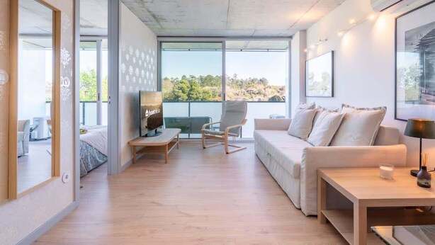 A modern living room with a sofa, a coffee table, and a TV. There is a large window offering a view of greenery outside. - Luxurious Soft Flat | Balcony and Pool by HostWise (Vila Nova de Gaia)