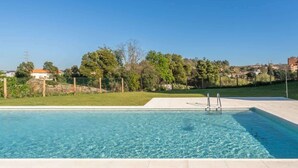 A rectangular swimming pool with clear blue water, surrounded by a tiled deck and a grassy area with trees and a fence. - Luxurious Soft Flat | Balcony and Pool by HostWise (Vila Nova de Gaia)
