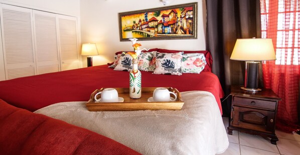 Una cama bien hecha con una colcha roja, una bandeja de madera con tazas y un florero, y un cuadro en la pared.