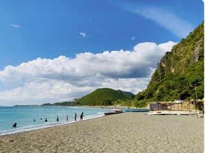 Una playa con gente nadando y caminando por la orilla, un bote pequeño y una ladera rocosa al fondo.