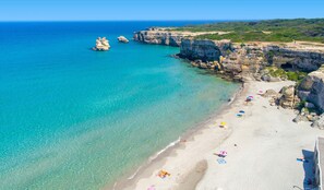 Ein Strand mit klarem türkisblauem Wasser, Felsküsten und einem Sandstrand mit Sonnenschirmen und Badegästen.